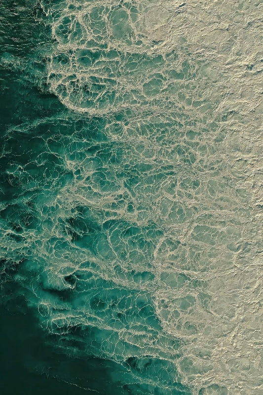 Aerial view of ocean water with swirling turquoise and deep blue sea meeting pale foam on the shore.