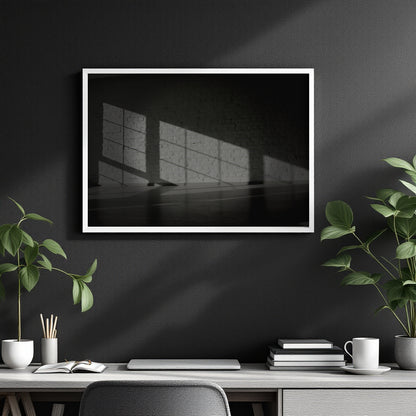 A framed black and white art print depicting light casting shadows on a wall, displayed on a dark gray wall above a modern desk with plants and books.