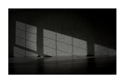 A black and white photograph of a brick wall with dramatic diagonal sunlight casting strong shadows across the wall and floor.