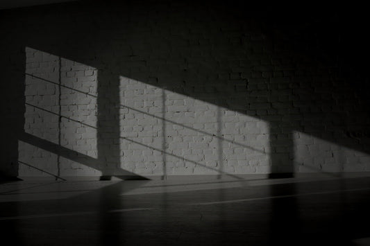 A moody black and white photograph of a white brick wall with dramatic, sharp window shadows cast across the surface and floor.