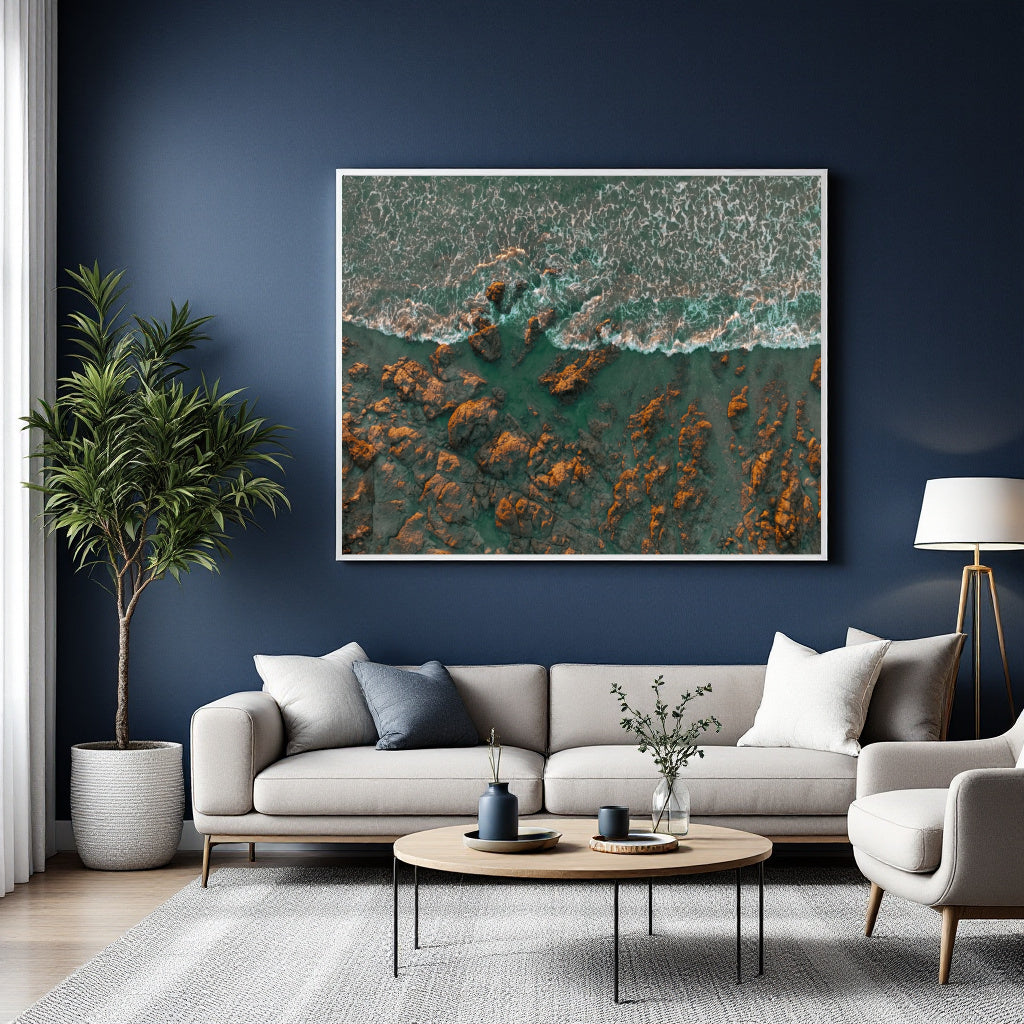 A large framed square wall art print showing an aerial view of turquoise water and orange-brown rocks, displayed above a light gray sofa in a modern living room with a dark blue wall.
