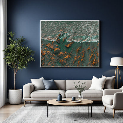 A large framed square wall art print showing an aerial view of turquoise water and orange-brown rocks, displayed above a light gray sofa in a modern living room with a dark blue wall.