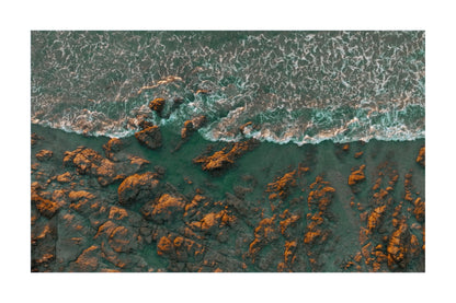 Aerial view of ocean waves with white foam crashing over dark rocky shoreline, with turquoise water and orange-brown rocks below, shown as a square print centered on a white background.