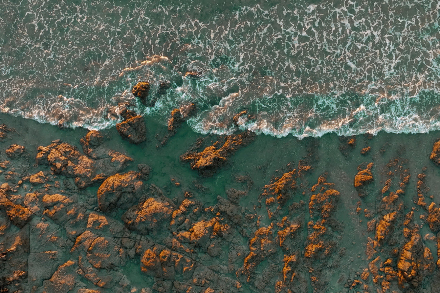 Aerial view of turquoise ocean waves crashing against a rocky shoreline with orange-brown rocks.