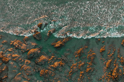 Aerial view of turquoise ocean waves crashing against a rocky shoreline with orange-brown rocks.
