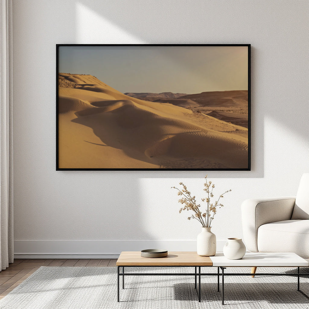 A large framed art print depicting golden desert sand dunes, displayed on a white wall in a modern living room setting above a low wooden coffee table.