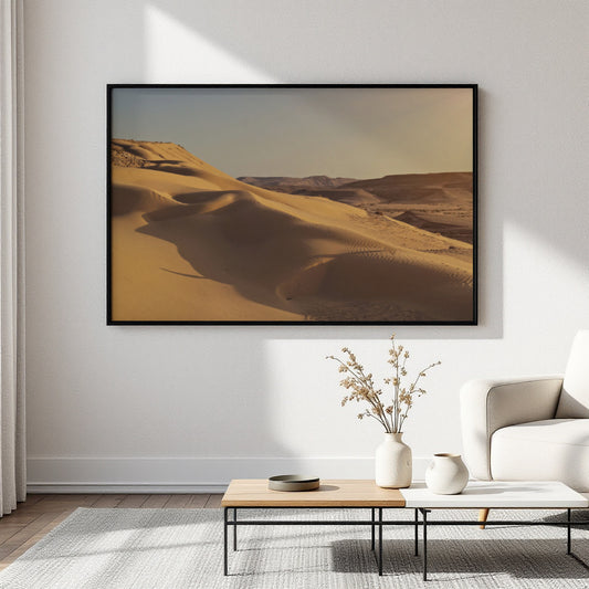 A large framed art print depicting golden desert sand dunes, displayed on a white wall in a modern living room setting above a low wooden coffee table.