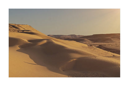 A scenic photograph of rolling sand dunes in a desert landscape under a pale sky, with warm golden light and soft shadows.