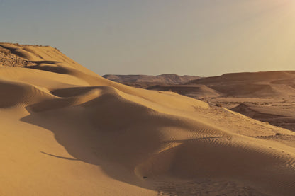 A scenic desert landscape with rolling sand dunes in warm golden-brown tones under a pale sky, with distant rocky hills in the background.