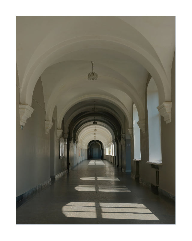 A long architectural corridor with repeating arched ceilings, pale walls, and sunlight streaming through windows onto the floor.