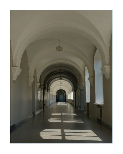 A long architectural corridor with repeating arched ceilings, pale walls, and sunlight streaming through windows onto the floor.