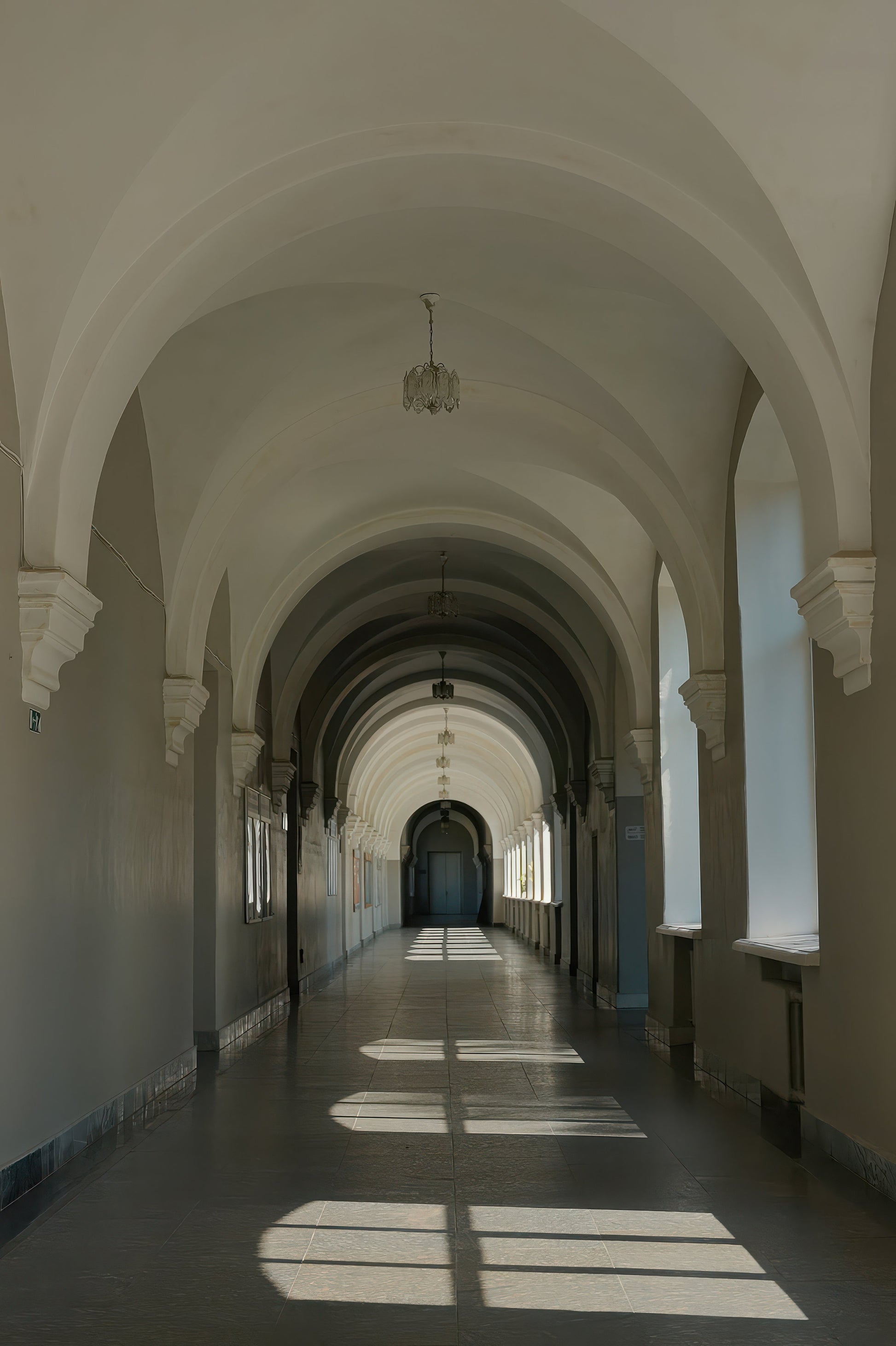 A long arched corridor with white arches and walls, a dark floor, and sunlight streaming in from windows on both sides.