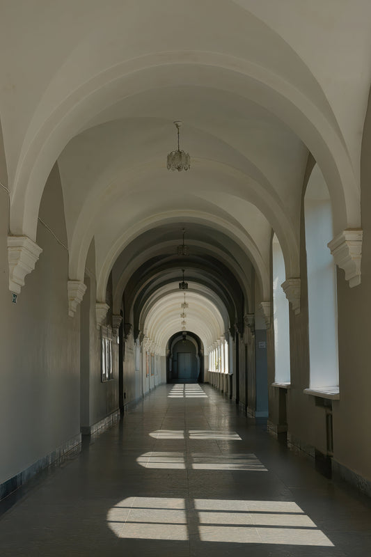A long arched corridor with white arches and walls, a dark floor, and sunlight streaming in from windows on both sides.
