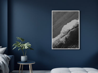 A vertical black and white framed art print of a crashing ocean wave, displayed on a dark navy blue wall in a modern bedroom setting next to a small plant on a side table.