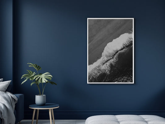 A vertical black and white framed art print of a crashing ocean wave, displayed on a dark navy blue wall in a modern bedroom setting next to a small plant on a side table.