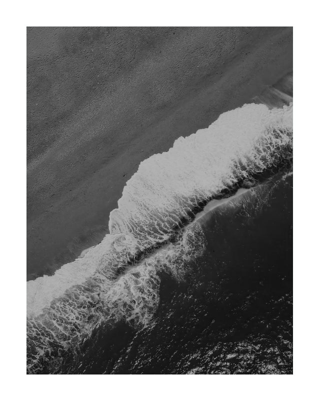 Black and white aerial photograph of ocean waves breaking onto a sandy beach, presented with a white border.