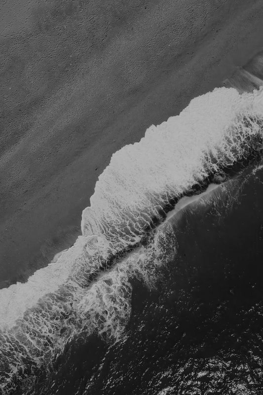 Black and white aerial photograph of ocean waves with white foam crashing onto a dark sandy beach.