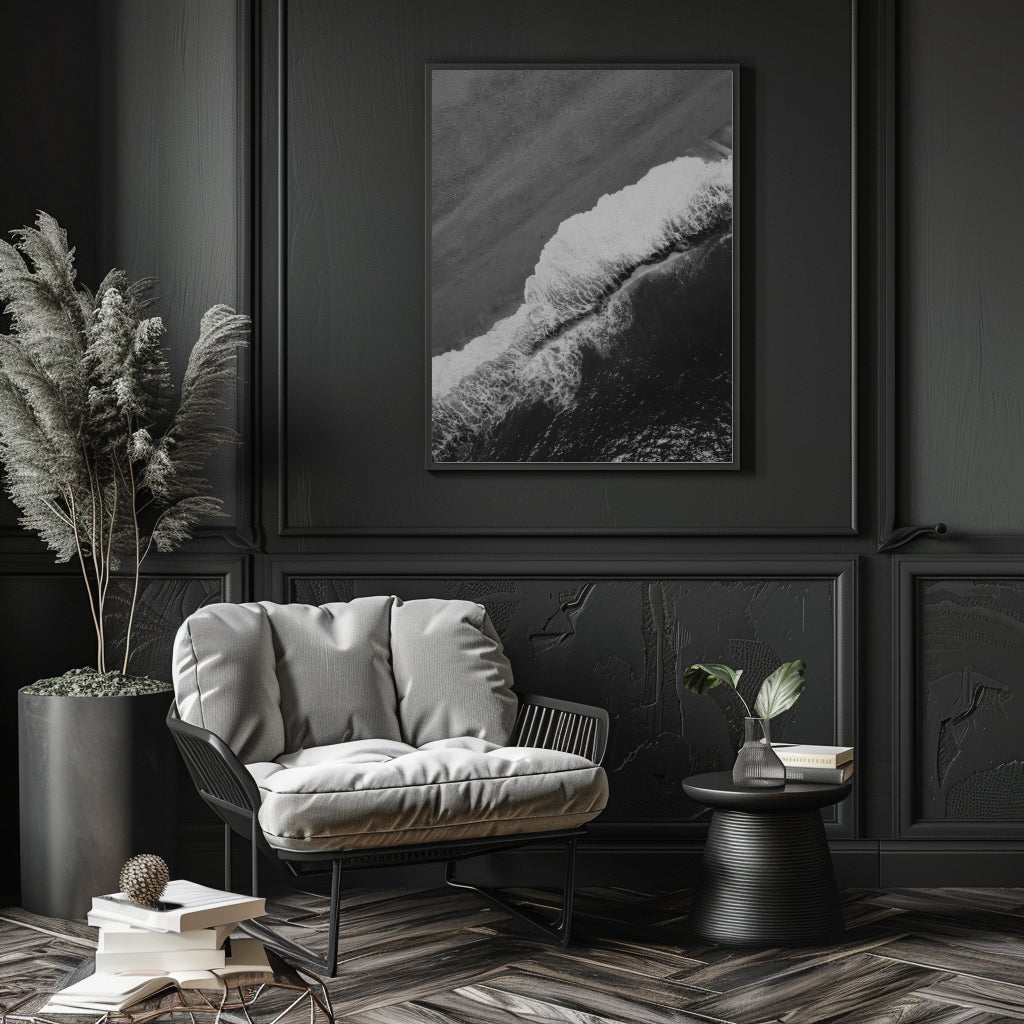 A black and white framed wall art print depicting a dramatic ocean wave, styled in a dark, elegant room above a gray lounge chair.