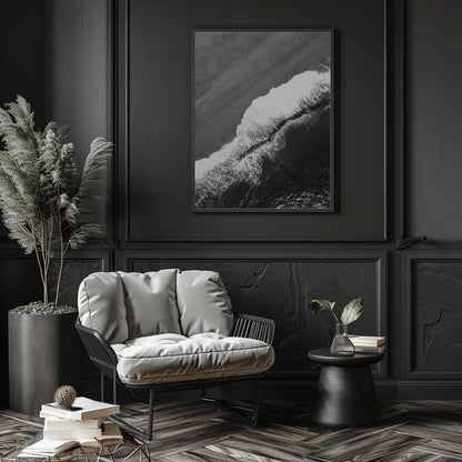 A black and white framed wall art print depicting a dramatic ocean wave, styled in a dark, elegant room above a gray lounge chair.