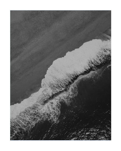 Black and white aerial photograph of ocean waves washing onto a sandy beach, presented as a square art print with a white border.