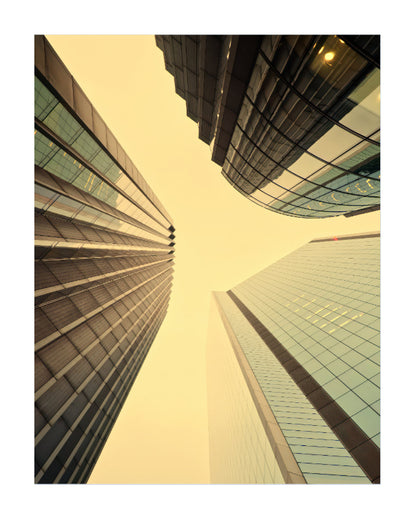 Low-angle vertical photograph of modern skyscrapers with glass curtain walls, viewed from below looking up at the buildings against a pale yellow sky.