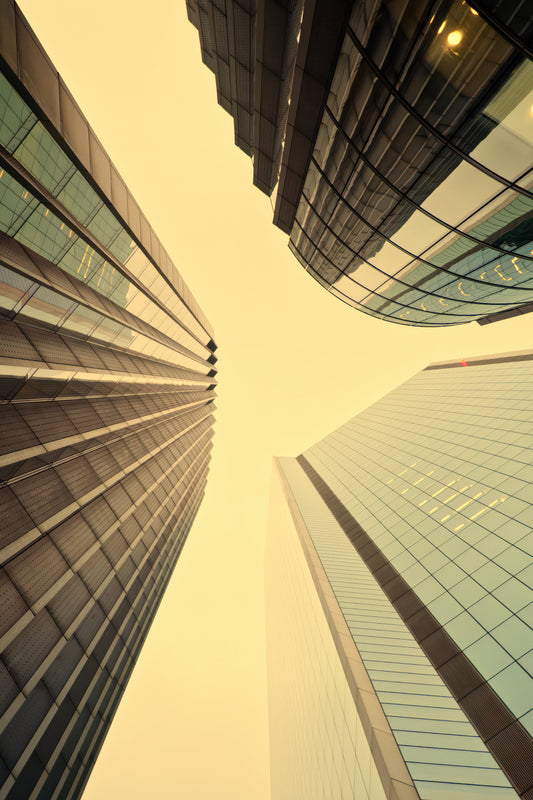 Low-angle vertical upward view of modern glass skyscrapers framing a pale yellow sky between the buildings.
