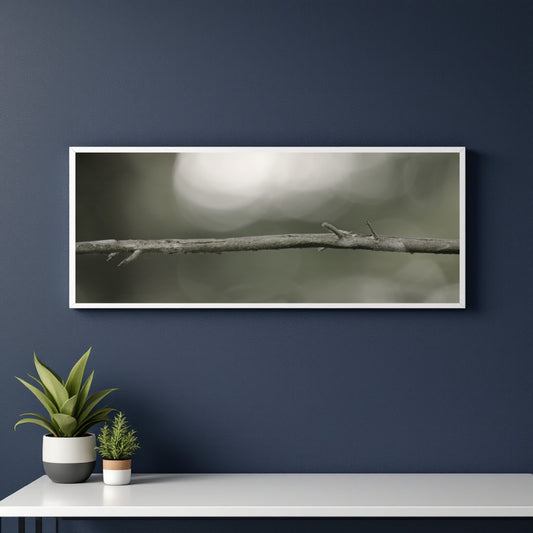 A horizontal framed art print featuring a close-up photograph of a thin tree branch against a soft, blurred gray-green background, displayed on a dark navy blue wall above a white console table with small potted plants.