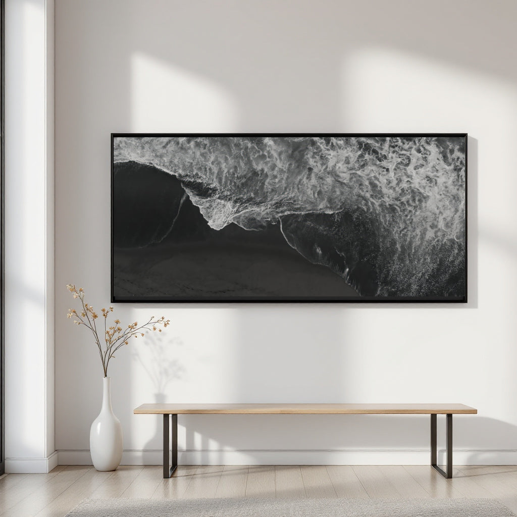A large black and white framed wall art print depicting ocean waves, displayed in a minimalist room setting above a light wood bench and white vase.