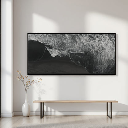 A large black and white framed wall art print depicting ocean waves, displayed in a minimalist room setting above a light wood bench and white vase.