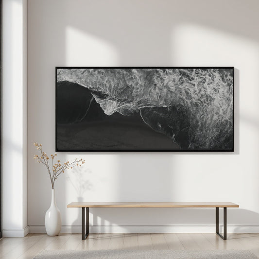 A large black and white framed wall art print depicting ocean waves, displayed in a minimalist room setting above a light wood bench and white vase.