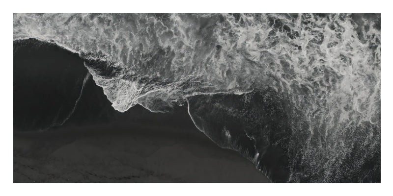 A black and white aerial photograph of a large ocean wave curling over dark sand or shallow water, with foamy white surf dominating the upper portion.