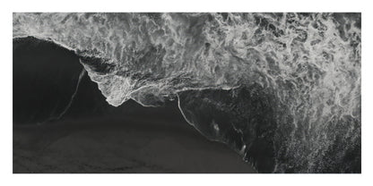 A black and white aerial photograph of a large ocean wave curling over dark sand or shallow water, with foamy white surf dominating the upper portion.