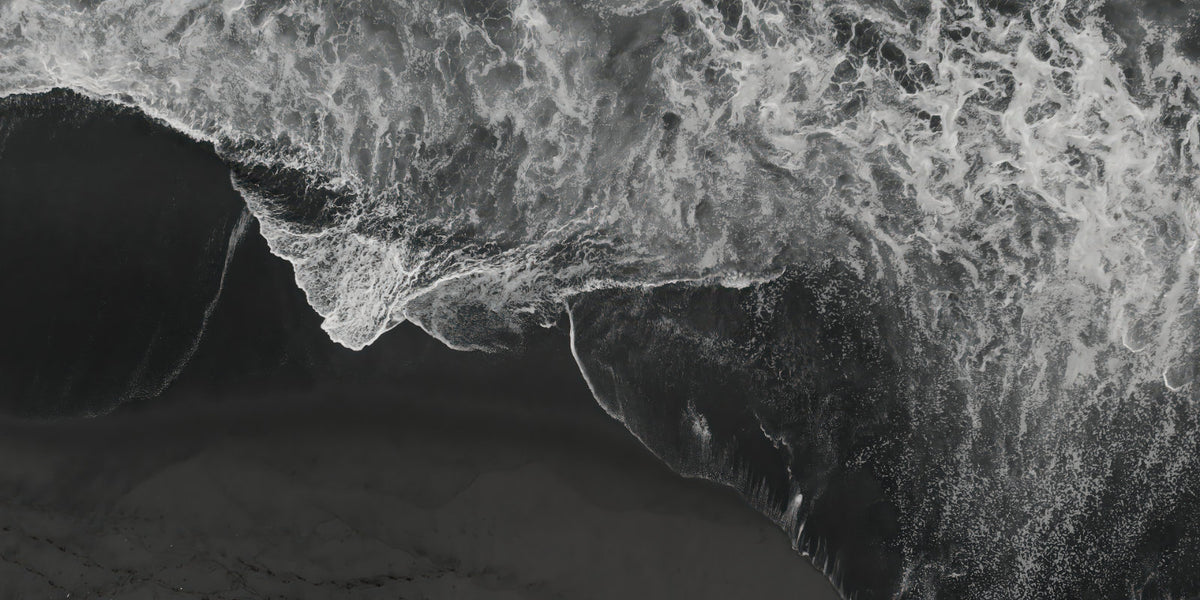 Black and white aerial photograph of a dramatic wave curling over a dark shoreline, with white foam contrasting against deep black and gray water.