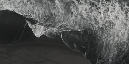 Black and white aerial photograph of a dramatic wave curling over a dark shoreline, with white foam contrasting against deep black and gray water.