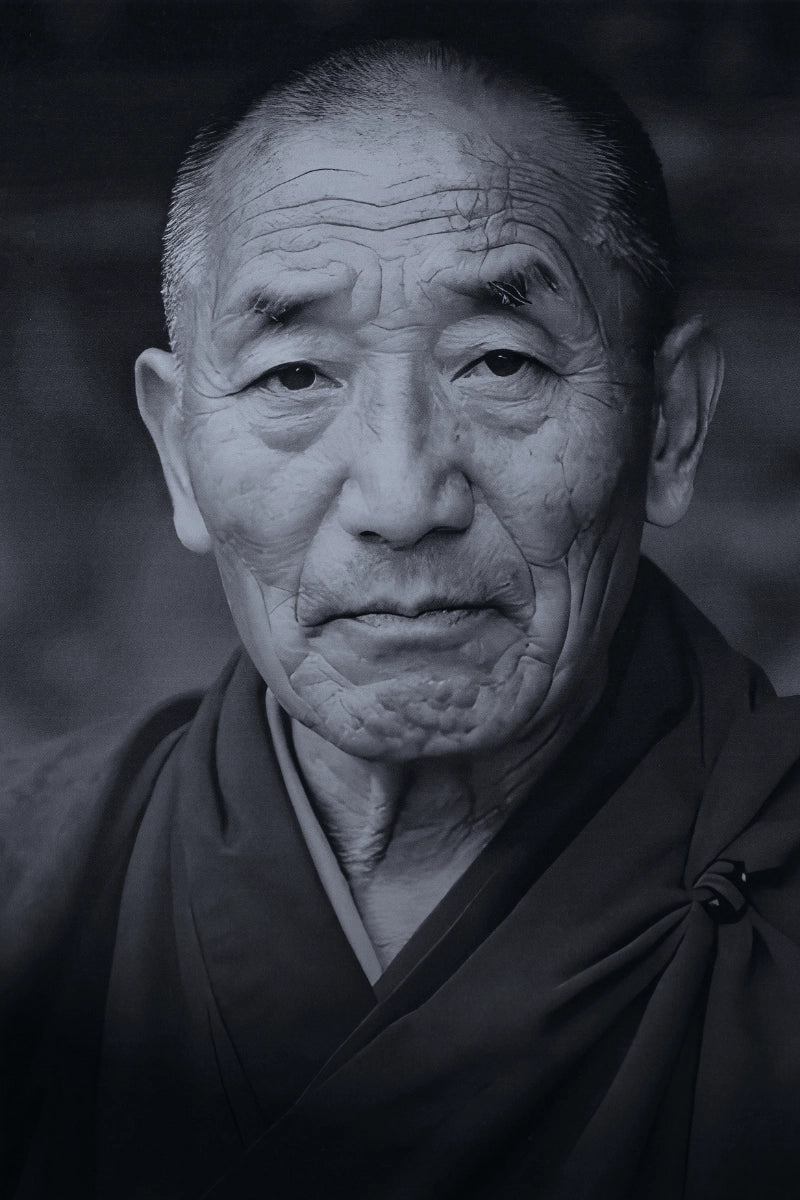 A black and white close-up portrait of an elderly man wearing dark robes, with detailed facial wrinkles and a blurred dark background.