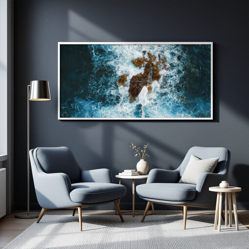 A large framed wall art print showing a dramatic aerial view of blue ocean waves crashing around brown rocks, displayed in a modern living room setting above two blue armchairs.