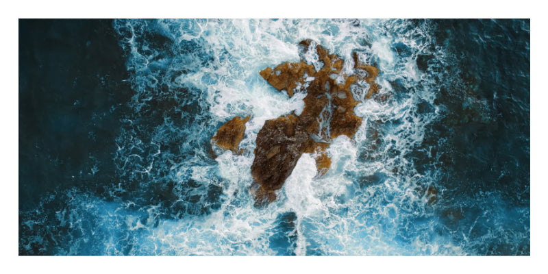 A square art print depicting an aerial view of dark blue ocean waves crashing around a rocky island, framed by a white border.