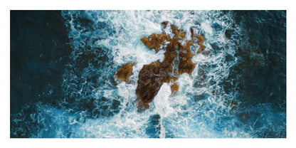 A square art print depicting an aerial view of dark blue ocean waves crashing around a rocky island, framed by a white border.