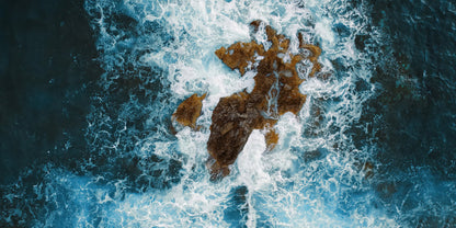 Aerial view of dark blue ocean water crashing around a rocky island, with white surf surrounding the brown and gray stones.