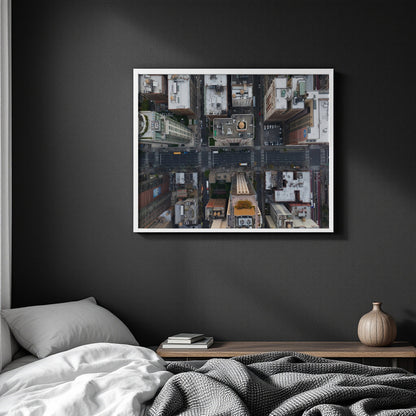 A framed square wall art print showing an aerial view of a city street grid with buildings, parked cars, and a yellow bus, displayed above a bed in a dark bedroom setting.