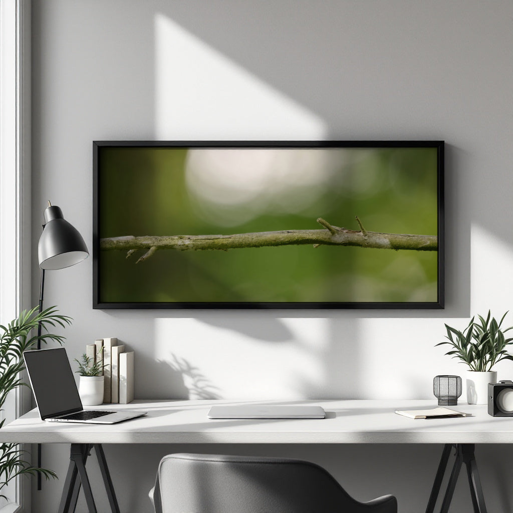 A framed wall art print showing a close-up of a green branch against a soft blurred background, displayed above a white desk in a modern home office setting.