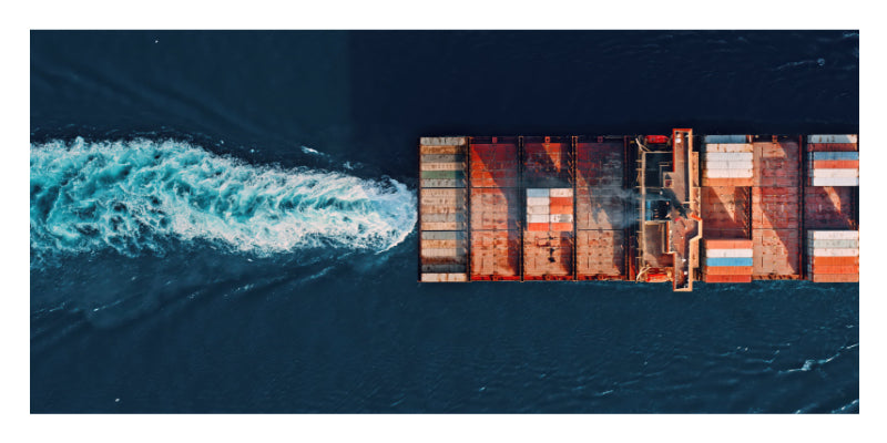 Aerial overhead view of a cargo ship carrying stacked shipping containers, leaving a white wake in dark blue water.