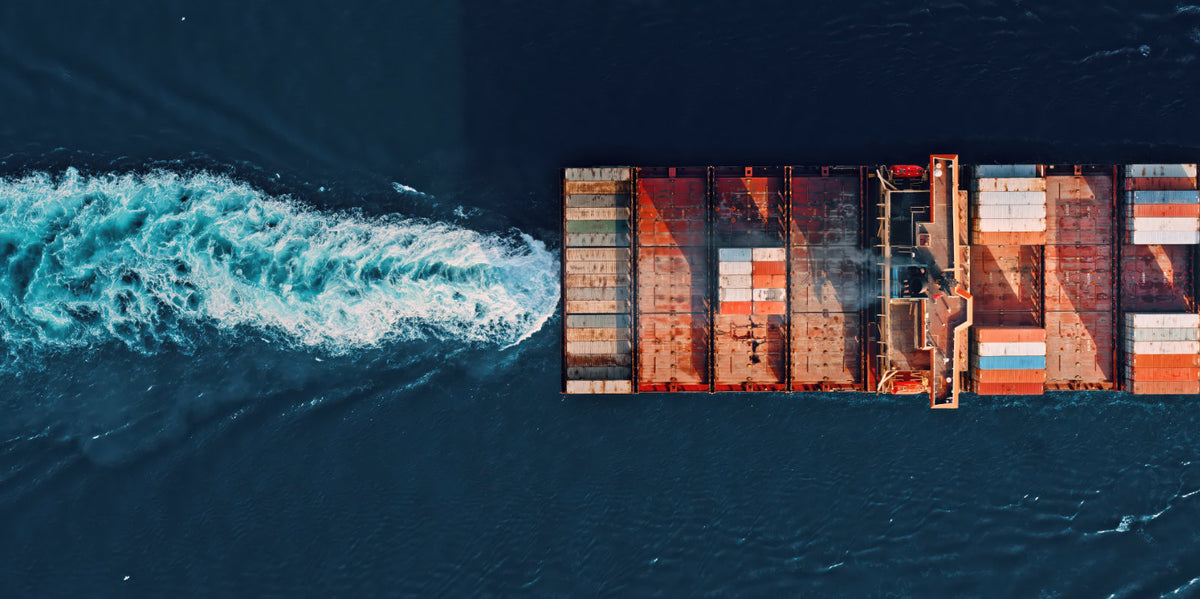Aerial view of a cargo ship carrying stacked shipping containers, moving through dark blue water and leaving a white wake.