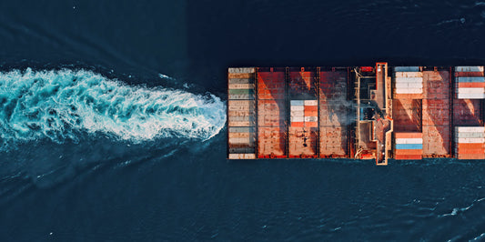Aerial view of a cargo ship carrying stacked shipping containers, moving through dark blue water and leaving a white wake.