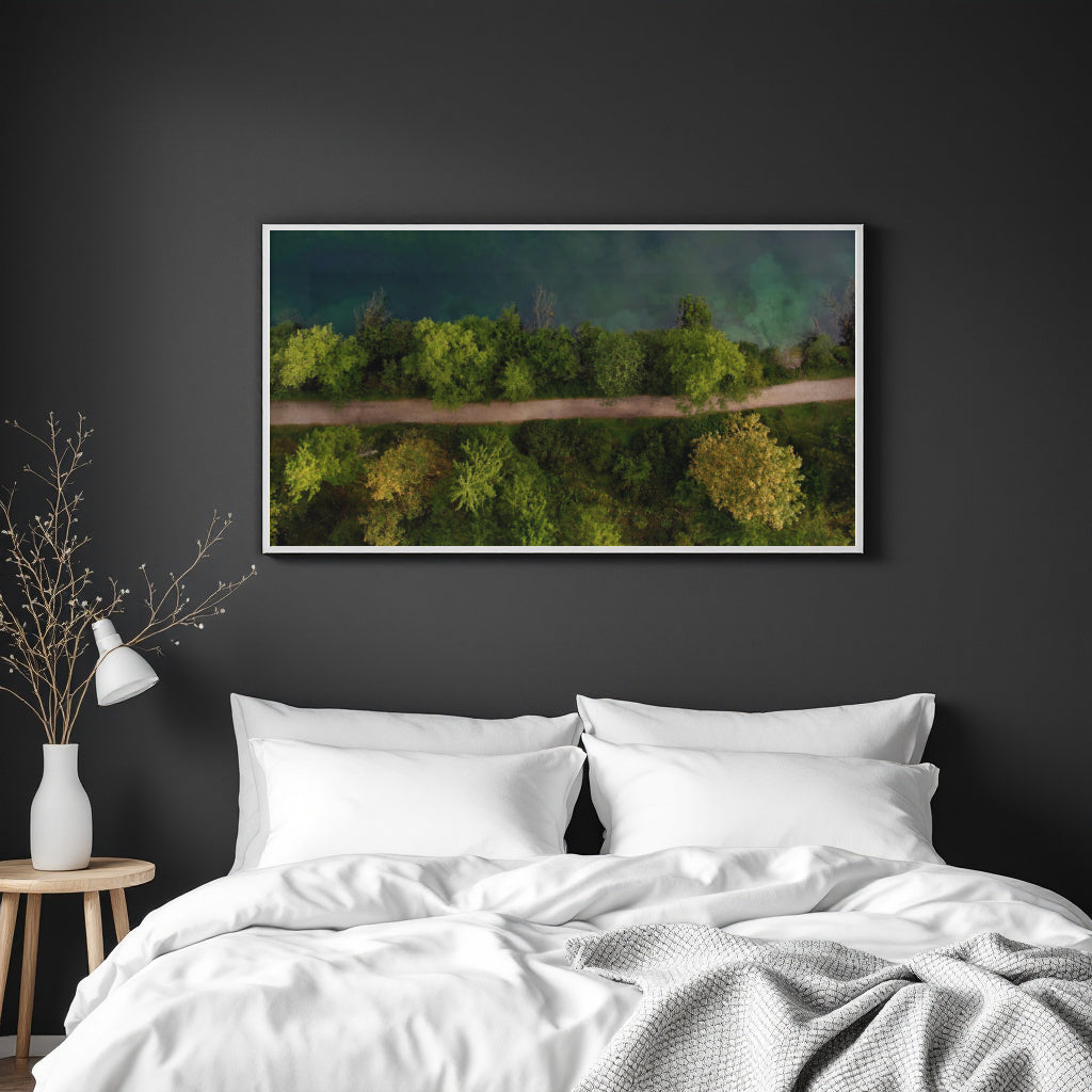 A framed landscape art print depicting a green forest and its reflection in water, hung on a dark gray wall above a neatly made bed with white bedding.