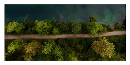 Aerial view of a narrow dirt road running horizontally through a dense green forest, with dark blue-green water visible along the top edge.