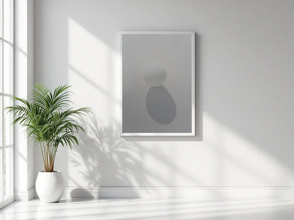 A minimalist framed wall art print with abstract gray shapes hangs on a white wall in a bright room with natural window light and a potted plant nearby.