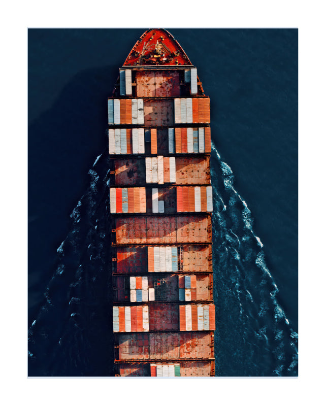 Aerial view of a cargo ship filled with stacked shipping containers, moving through dark blue water and leaving a white wake.