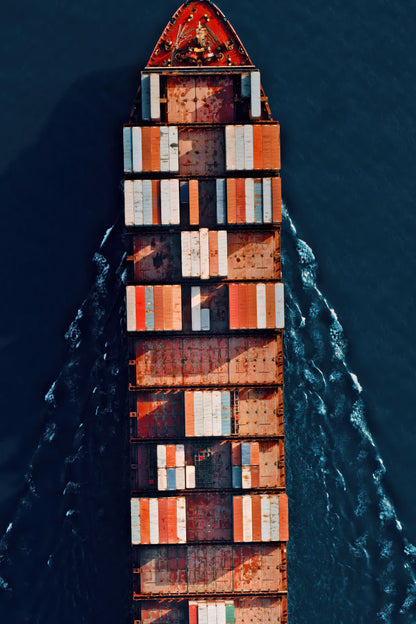 Aerial view of a large cargo ship packed with stacked shipping containers, moving through dark blue water and leaving a white wake.