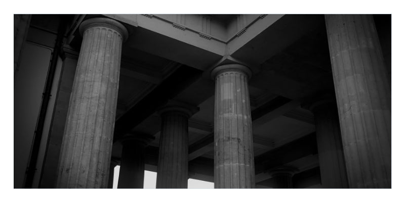 Black and white architectural photograph of massive classical columns supporting a grand structure, with dramatic high-contrast lighting.